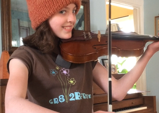 Young girl, Ella Thompson Rose playing the violin as she smiles into the camera, wearing a burnt orange knit hat and a piano in the background.